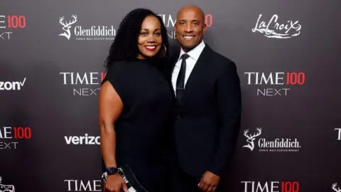 Getty Images Victor Glover stands on the right in a dark suit, white shirt and slim black tie, his arm gently around Dionna’s back as they pose together on the red carpet. Dionna, on the left, wears a simple knee‑length black dress and carries a patterned clutch bag in her right hand. They stand close, facing the camera against a dark backdrop printed with “TIME100 Next” and sponsor logos, giving a formal but relaxed, celebratory feel