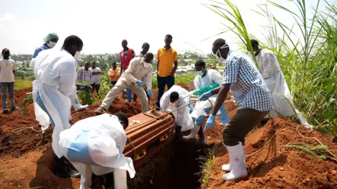 Amensisa Negera / BBC People wearing protective gear, such as masks and gowns, lower a coffin into the ground using ropes