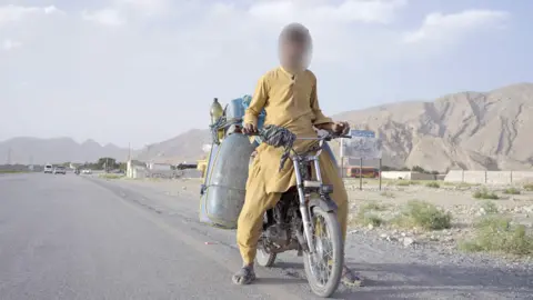 Mazaar, who is dressed in yellow, flowing shirt and trousers, is pictured on his motorbike with huge cannisters of fool strapped to the back. He's on a road with mountains in the background.