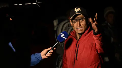 Luis ACOSTA / AFP via Getty Images The relative of a miner caught in the explosion at the mine in Sutatausa gestures as he speaks to the media. His face is stained with coal dust. He is wearing a black baseball cap with a white cockerel embroidered on top and a red coat. 