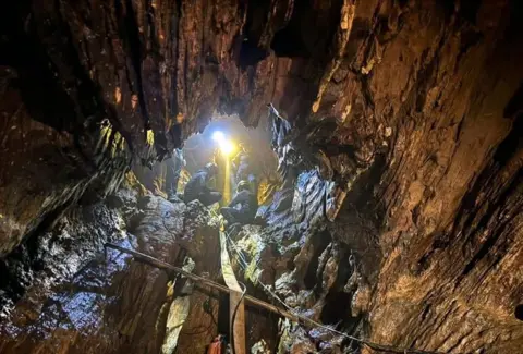 EPA An enormous rocky cavern fills the image with the centre showing the entrance to the cave and four rescuers dressed in blue outfits and helmets using a yellow pump hose, which stretches all the way to the bottom of the picture. A bright yellow light picks out the details of the craggy rocks inside the cave from where they are standing