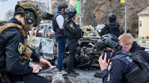 EPA Six Ukrainian experts wearing helmets, and some of them wearing dark blue uniforms, work at the site of a drone attack in the Ukrainian city of Kharkiv. Behind them stands a wrecked car. 