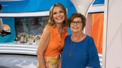 NBC via Getty Images Savannah Guthrie and mother Nancy Guthrie on the Today show set in June 2023. Savannah has long light brown hair and is wearing an orange sleeveless shirt and colourful floral trousers. Nancy has short brown hair and is wearing a blue top with a long necklace