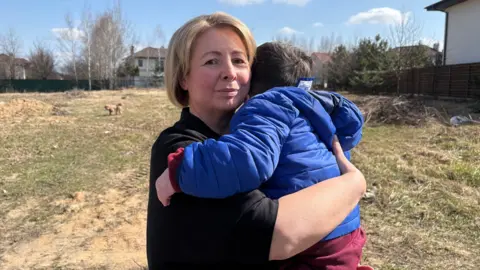 A woman holds a young child in her arms. The boy’s back is turned to the camera so his face cannot be seen. He has dark hair and is wearing a blue jacket and red trousers. The woman is looking at the camera – she has short, straight, blonde hair and is wearing a black top. They are outside and rough grass and a handful of houses can be seen behind them