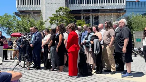BBC News At a distance, a man stands at a mic with a group of people surrounding him. they are in front oiif a courthouse outside with blue sky behind them