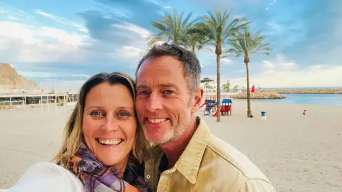 Free Lindsay and Craig Campaign A couple on a beach. It is sunny and the sand is white. Behind them are some trees.