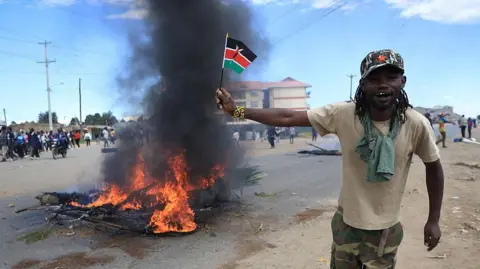 Anadolu via Getty Images A protester holds a small Kenyan flag along a road with a fire burning and groups of protesters gathered in the background