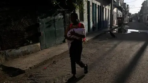 REUTERS/Norlys Perez A woman walks with a baby as Cuba begins efforts to restore power after its grid collapsed in Havana, Cuba on 22 March, 2026. 