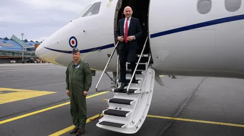 Getty Images John Healey in a white shirt, black suit and red tie, seen exiting an RAF plane at an airport as a soldier stands to attention nearby