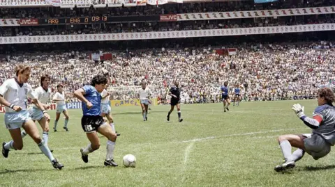 AFP via Getty Images Maradona is seen dribbling the ball through a number of English footballers and the English goalkeeper is seen diving to the side.