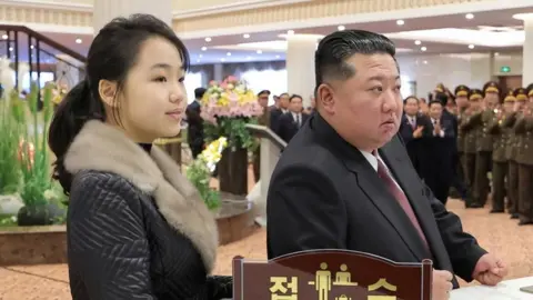AFP via Getty Images With her long hair tied back over the light grey fur collar of a dark grey jacket, Kim Ju Ae, stands next to Kim Jong Un, in a matching suit, white shirt and burgundy tie, inside the lobby of the Milyong Hotel, in the Samjiyon tourist district of Ryanggang Province. In the background, several rows of besuited and uniformed dignitaries applaud, alongside colourful bouquets, lighting and a staircase.
