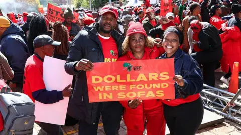 Gallo Images via Getty Images Three EFF supporters dressed in red hold up a sign that says 'dollars were found in sofas'. They are standing in front of a large crowd of EFF supporters.