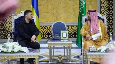 Reuters Ukraine's President Volodymyr Zelenskiy meets Deputy Governor of Makkah Region Prince Saud bin Mishaal bin Abdulaziz. The pair are seated in elaborate chairs with a silver-and-gold table between them. The room they are in is palatial, with a gold-and-black printed pattern on the walls. Zelensky is wearing an all-black outfit with a jacket, and the Deputy Governor wears a red-and-white traditional headscarf.