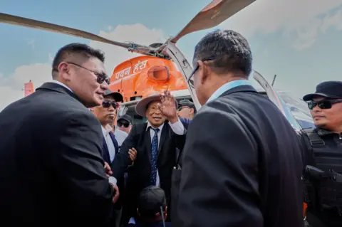 AP Dressed in a black coat, white short and blue tie, Thuingaleng Muivah waves at his supporters after getting out of the helicopter that brought him back home