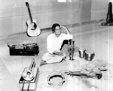 Stills Ravi Ilaiyaraaja sits next to musical instruments including a violin, trumpet, guitar, bongos saxophone