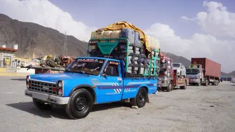 A blue pick-up truck carrying cannisters of oil. Behind it are othr vehicles and in the background the mountains