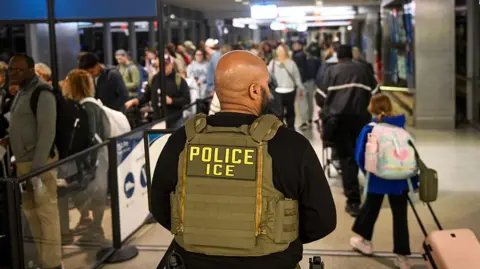 Getty Images The back of a male ICE agent wearing a vest that reads police and ice below it, in capital letters. He is standing in an airport - behind him are lots of people, some with luggage.