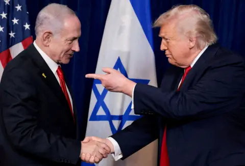 Reuters Benjamin Netanyahu and Donald Trump shake hands while standing in front of US and Israeli flags. Both men are wearing dark blue suits with red ties, while Trump is pointing at Netanyahu.