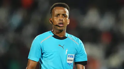 Getty Images Omar Artan, wearing a blue referee's top bearing a badge reading 'Fifa Referee 2025' looks towards the camera while he is officiating a football match