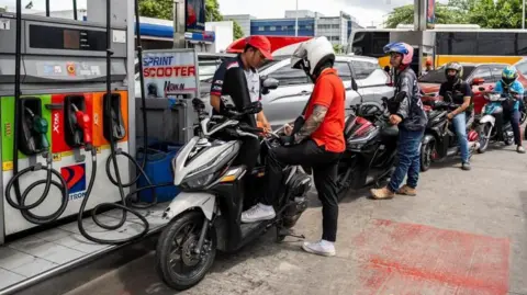 Reuters A worker helps a motorcyclist top up fuel for his bike. Behind him are more than five other bikers queuing for fuel.