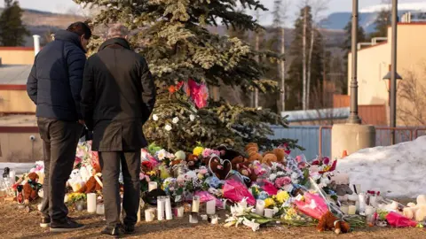 AFP via Getty Images Two men standing outside, backs to camera looking down at a large collection of flowers and presents left for the victims of the school shooting, placed underneath a large tree.