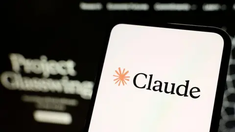 NurPhoto via Getty Images A smartphone screen displaying the name Claude and a drawing of an asterisk. Out of focus in the background the words Project Glasswing are legible.