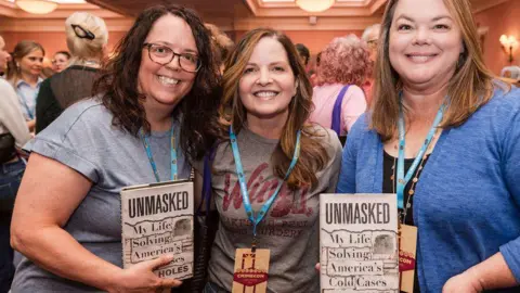 CrimeCon and RenownedPhotos Three female attendees wearing lanyards hold up books titled: "Unmasked - My Life solving America's Cold Cases"