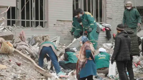 BBC About six people in green jackets look through the rubble outside a building. The remains of two windows with vertical white bars across the holes can be seen. Another three men are looking on and a woman walks past.