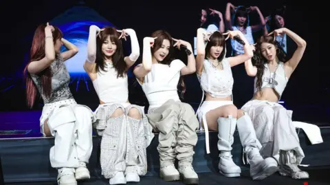 Source Music Le Sserafim sit on stage during their EasyCrazyHot tour, making 'cat ears' with their fingers, while wearing toned grey and white street wear