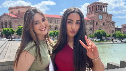 Two young women with long hair stand side by side in front of a brick building with a red roof. The woman on the left smiles at the camera, while the one of the right pouts and puts her hand up in a peace symbol.