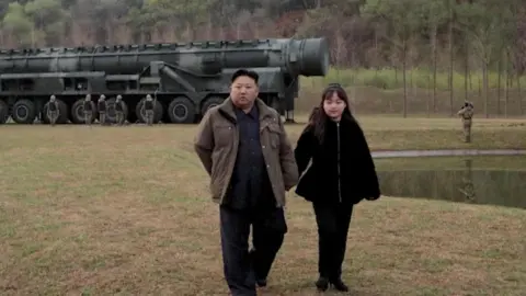 Reuters Kim Jong Un, in a black suit and open brown coat, and Kim Ju Ae, with her long hair tied back, in a $1,900 Christian Dior black padded jacket and trousers, walk on grass beside a lake, in front of a Hwasong-18 intercontinental ballistic missile (ICBM), with several men standing next to it, and trees in the background. 