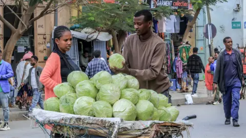 AFP via Getty Images A male vendor holds up one of many cabbages he is selling for a woman in a black T-shirt and pink cardigan on a street in Tigray, Ethiopia - March 2026