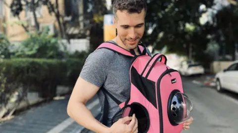 Tom Dan, looking down on the ground and smiling as he wears a grey T-shirt, and a pink backpack on his front, in the middle of a road where some white cars are parked to the right and a green hedge at the edge of the pavement on the left, and green trees in the background