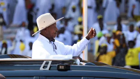 Reuters Uganda's President Yoweri Museveni, in a white shirt and a white hat, salutes his supporters from an open-top black vehicle after his swearing-in ceremony - 12 May 2026