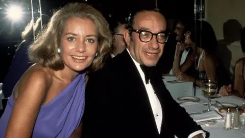 Archive Photos via Getty Images Barbara Walters and Alan Greenspan are photographed at a black tie dinner in 1977. They are looking at the camera and smiling. Waters is wearing a blue dress and Greenspan is wearing a tuxedo