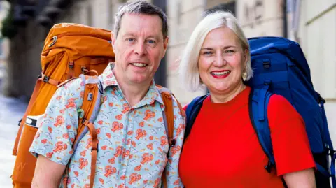 BBC Mark Blythen, in his 60s, who has short grey hair and is wearing a short sleeved blue shirt with a lobster pattern and a large orange backpack, stands next to Margo Oakley, who has short blonde hair, a red t-shirt and a large blue backpack. 