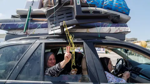 Reuters A displaced Lebanese woman smiles and gestures as she sits inside a car with her relatives during their journey home, on the highway near Sidon, southern Lebanon (16 June 2026)
