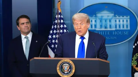 Getty Images Trump is at the podium and speaking while wearing a dark blue suit and bright blue tie. To his right and behind him is Todd Blanche who is wearing a dark suit and indigo tie