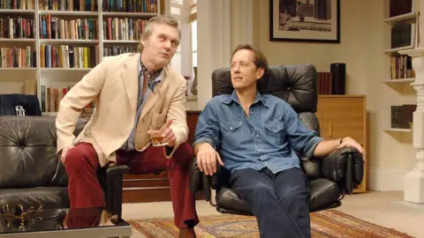 PA Media Head and Richard E Grant sit next to each other in character on set - the set is a living room, and Head holds what appears to be an alcoholic drink in his hand