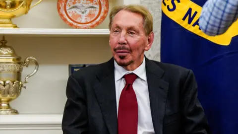 Reuters Oracle co-founder Larry Ellison sitting in a chair at the White House wearing a dark grey suit, white dress shirt and deep red neck tie.