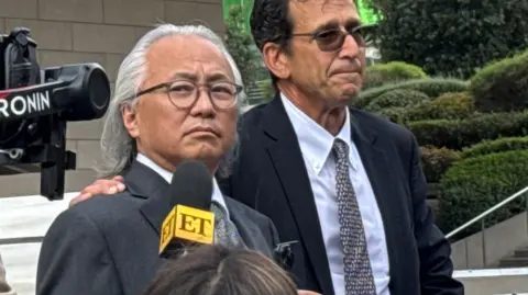 Kenneth Iwamasa, in a grey suit and tie and light rimmed glasses, looks to the right stands next to his lawyer, who has his hands around his shoulder. A media camera and a microphone are in the frame.