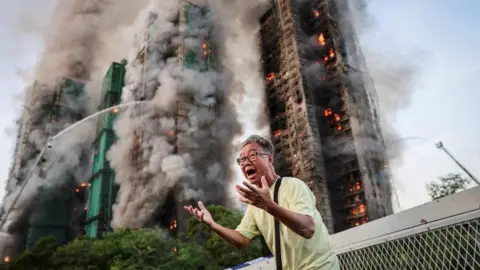 Reuters A man holds on both hands, shouting in agitation, as long columns of smoke billow out from the residential blocks in the background. Some units continue to burn even as firefighting has started