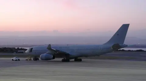 PA Media A Voyager aircraft - which resembles a passenger airliner and is grey with 'Royal Air Force' written on the side in a darker grey - sits on the runway at RAF Akrotiri. The sun is setting in the background.