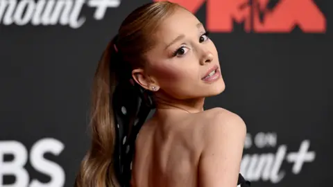 Noam Galai/Getty Images for MTV Ariana Grande looks over her shoulder on the red carpet of the MTV awards. She wears her long auburn hair in a high pony tail.