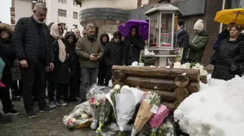 EPA Relatives of victims observe a minute of silence around bouquets of flowers leaning on logs, with a candle-holding container on top, snow around, and with two women with their umbrellas unfurled.
