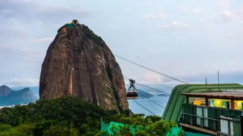 Emin Sansar/Anadolu via Getty Images A view of Sugar Loaf cable car at Guanabara Bay in November 2024. The steep rocky mountain can be seen against the blue of the bay. At the top of the mountain, the cable car station is visible. Cables connect it with Morro da Urca, from which the photo is taken. A cable car can be seen suspended halfway between the two. 