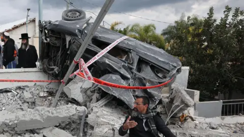 EPA Man sitting beneath wrecked car gestures with his hand