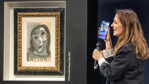 EPA A blue painting by Picasso, in his trademark cubist style, framed on the wall beside a woman holding a microphone up to her mobile phone, which shows a video call with a man wearing glasses - both are smiling broadly.