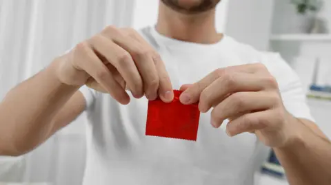 Getty Images A bearded man wearing a white t-shirt is seen opening a red condom packet with both of his hands inside a white coloured bedroom.
