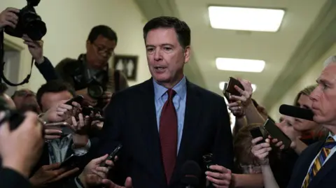 Getty Images James Comey is surrounded by reporters holding phones and mics and cameras as he appears in Congress to testify in 2018. 